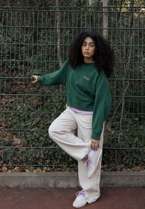 Γυναίκα με σγουρά μαλλιά που φοράει πράσινο φούτερ και μπεζ παντελόνι τεντώνεται όρθια σε ένα πόδι κρατώντας μεταλλική περίφραξη σε εξωτερικό χώρο.