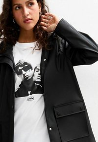 Czarna wodoodporna kurtka z zamkiem z przodu, dużą kieszenią i regulowanym kapturem, noszona na białej koszulce z nadrukiem graficznym w czerni.