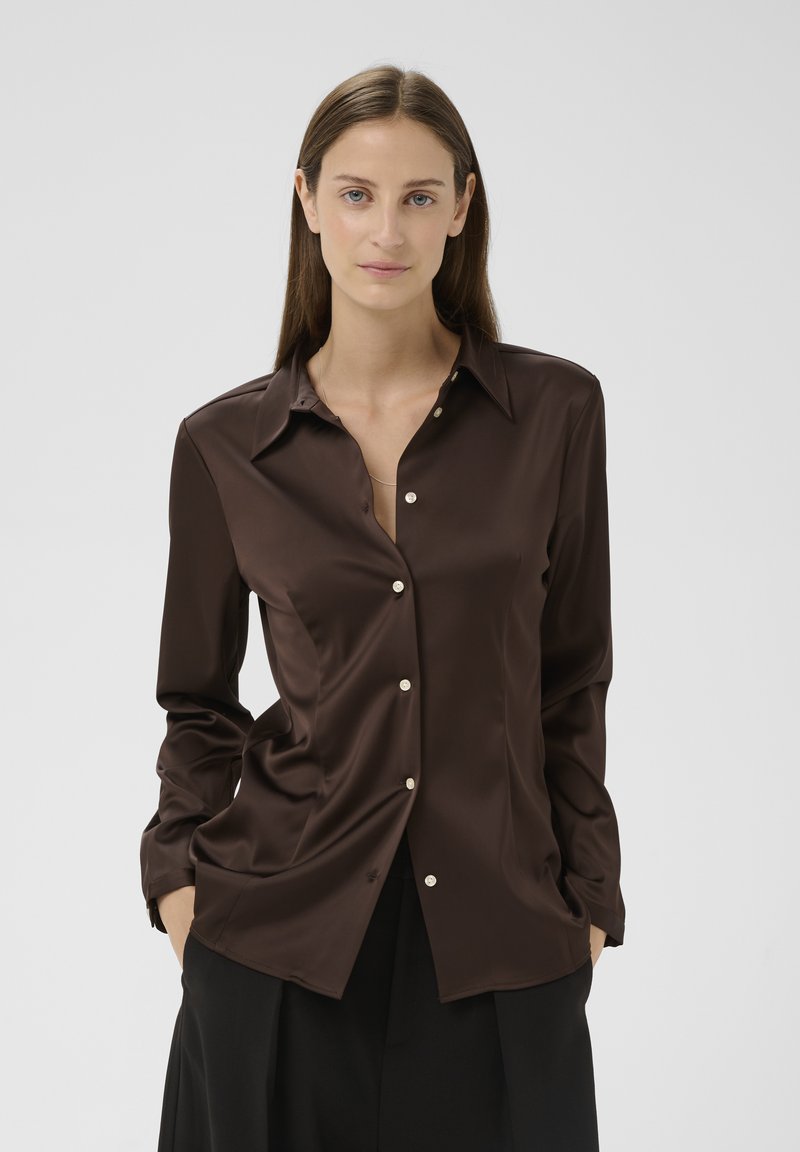 Femme portant un chemisier en satin brun foncé à boutons avec manches longues et un pantalon noir, debout les mains dans les poches devant un fond uni.