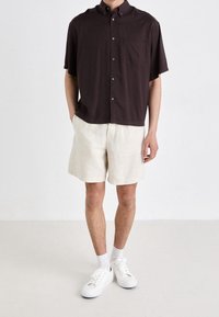 Kortbruin overhemd met knopen en een borstzak, gecombineerd met lichtbeige linnen shorts en witte sneakers. Soepele stoftextuur.