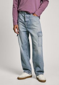 Pantalones cargo de mezclilla azul claro con dos bolsillos laterales, corte relajado y costuras visibles. Combinados con zapatillas blancas y una camisa de manga larga morada.