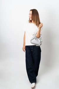 Kvinna i vit T-shirt och mörka jeans som håller en ljusgrå handväska, stående mot en enkel vit bakgrund, och tittar åt vänster.