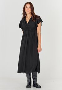 Svart maxiklänning med vingspetsärmar, markerad midja och flytande tyg; kombinerad med höga svarta stövlar för kontrast. Mjuk textur.