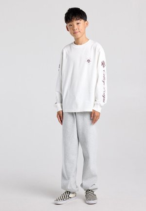 Jonge jongen staat draagt een wit shirt met lange mouwen met tekst op de mouwen, grijze joggingbroek en zwart-witte geruite slip-on schoenen.