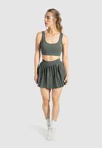 Donkergroene sportbeha met bijpassende geplooide rok. De stof is glad met een elastische tailleband. Witte sportschoenen en sokken maken de look compleet.