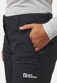 Hand mit hellrosa Nagellack in der vorderen Reißverschlusstasche der schwarzen Jack Wolfskin Hose, getragen mit einem weißen Langarmshirt.