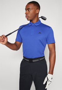 Blå kortermet polo med krage, svart logo, kombinert med mørke bukser og belte med svarte striper, holder en golfkølle.