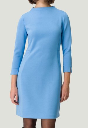 Femme portant une robe simple bleu clair, longueur genou, manches trois-quarts, avec un col rond haut, sur un fond uni.