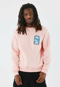 Sudadera de color rosa claro con puños y dobladillo acanalados, que presenta un parche gráfico de un diseño escénico en el área del lado izquierdo del pecho.