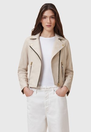 Beige Lederbikerjacke mit asymmetrischem Reißverschluss, goldenen Hardware-Akzenten und Reverskragen, getragen über einem weißen Shirt und hellen Jeans.