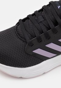 Schwarzer Sportschuh mit einem strukturierten Mesh-Obermaterial, lila Akzenten an der Seite und schwarzen Schnürsenkeln. Verfügt über eine weiße, gepolsterte Sohle.