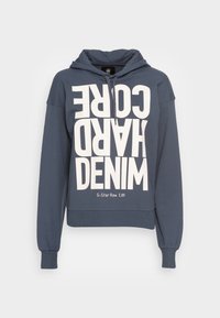 Blå hoodie med stor vit text i block "G-STAR RAW HARD CORE DENIM." Har dragsnodd i huvan och stickade cuffs. Slät tygtextur.