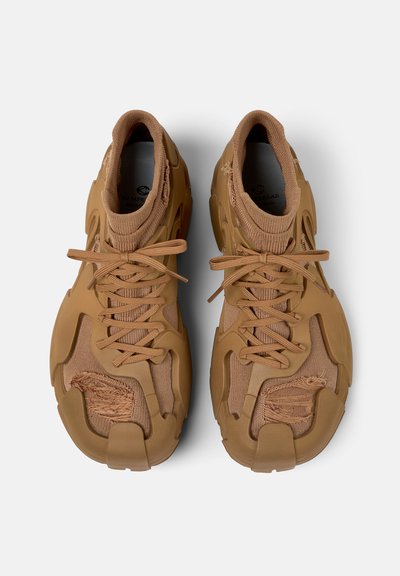 Paar hellbraune Sneakers aus Strickstoff mit klobiger Gummisohle und abgenutzten, zerrissenen Details im Zehenbereich, von oben betrachtet.