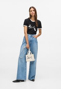 Mulher a usar uma t-shirt gráfica preta e jeans de perna larga azuis, segurando uma pequena mala bege com um design correspondente, de pé contra um fundo branco.