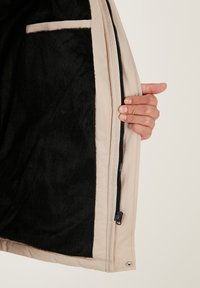 Țesătură exterioară de culoare bej, cu un interior din fleece negru texturat. Include un fermoar, un material pentru prinderea cu mâna și o buzunar lateral vizibil.