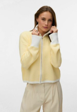 Kvinde justerer kraven på en lysegul trøje med lynlås og hvide manchetter, kombineret med beige bukser med høj talje, foran en ensfarvet baggrund.