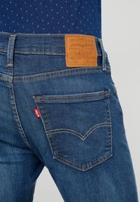 Blå denimjeans med en brun läderlapp i midjan, femficksdesign och en röd etikett på vänster bakficka.