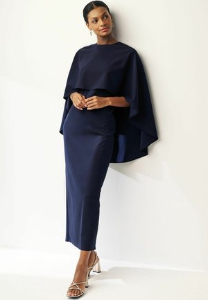 Navy gown with a draped, cape-like overlay, smooth texture, fitted silhouette, and paired with metallic sandals featuring block heels.