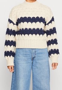 Paksu neulepusero, jossa on kerma- ja laivastonsinisiä sahalaitaisia raitoja, yhdistettynä vaaleansinisiin korkeavyötäröisiin laukkoihin, seisovalla henkilöllä.