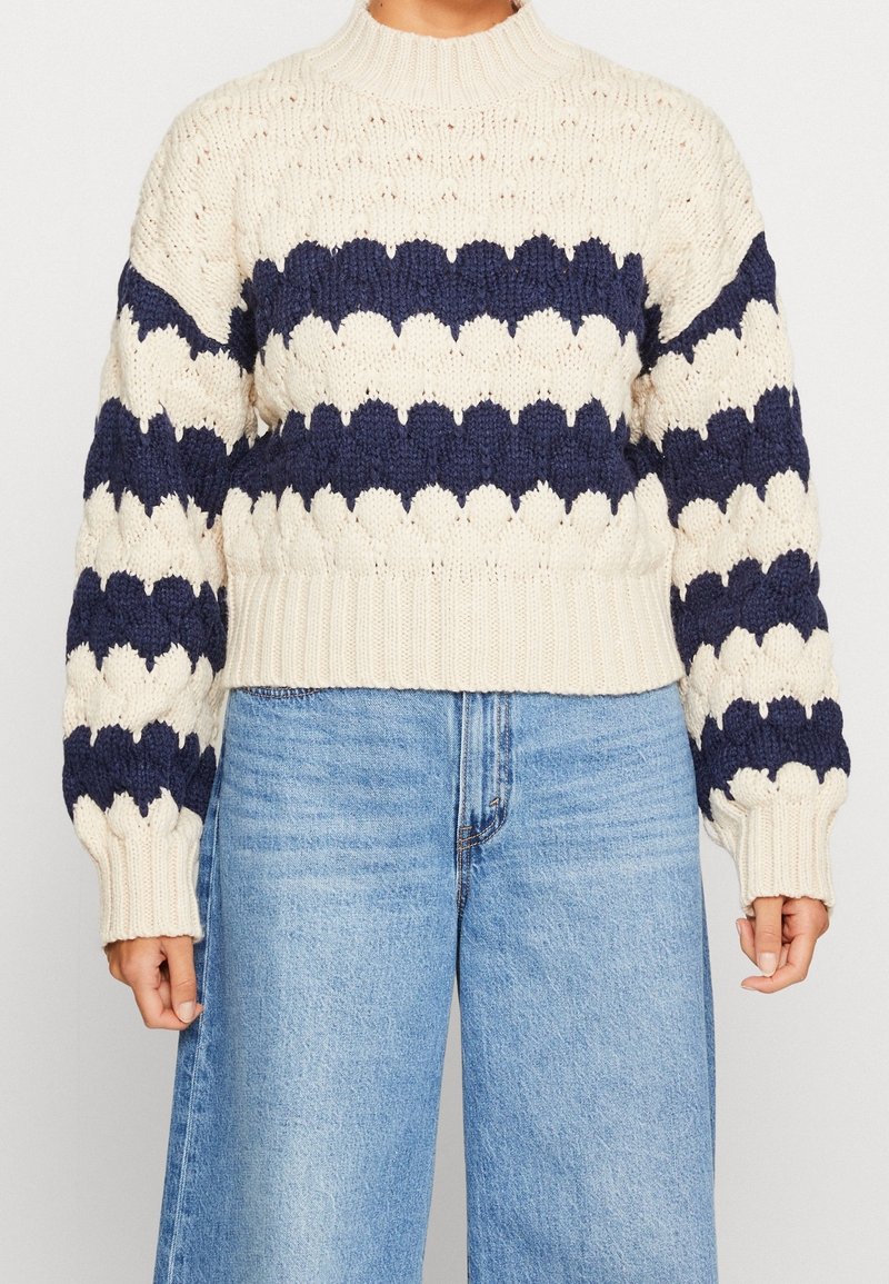 Paksu neulepusero, jossa on kerma- ja laivastonsinisiä sahalaitaisia raitoja, yhdistettynä vaaleansinisiin korkeavyötäröisiin laukkoihin, seisovalla henkilöllä.