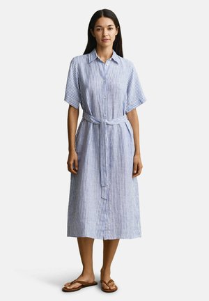 Femme debout portant une robe chemise midi à rayures verticales bleu et blanc, à manches courtes, avec une ceinture nouée à la taille et des tongs marron.