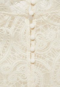 Vêtement en dentelle blanche présentant un motif floral, six boutons ronds recouverts de tissu et une texture translucide le long du centre.