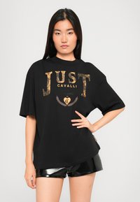Black oversized t-shirt with bold gold "JUST CAVALLI" logo. Features short sleeves and a round neckline, paired with shiny black shorts.