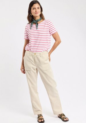 Femme debout, main dans la poche, portant un t-shirt blanc à rayures rouges, un pantalon beige, un foulard vert autour du cou et des sandales vertes, sur fond blanc.
