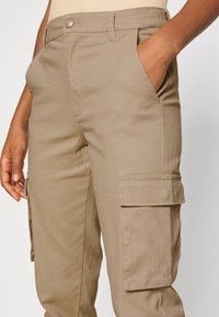 Pantalons cargo kaki avec une texture lisse, présentant une poche latérale et une poche avant. La taille est dotée d'un bouton en métal et de passants de ceinture.