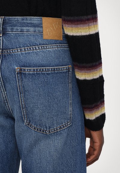 Jeans din denim albastru cu un finisaj texturat, având o pată din piele deschisă la nuanță la talie și un buzunar din spate cu detalii vizibile de cusătură.
