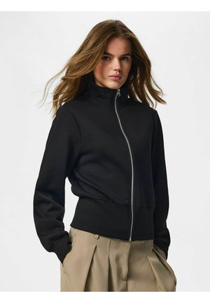Jonge vrouw met los bruin haar draagt een zwart jack met rits en beige geplooide broek met handen in de zakken tegen een effen achtergrond.