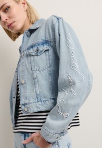 Lys blå denimjakke med en kort design, som har to lommer foran og hvite broderte detaljer på ermene. Stripete skjorte under.