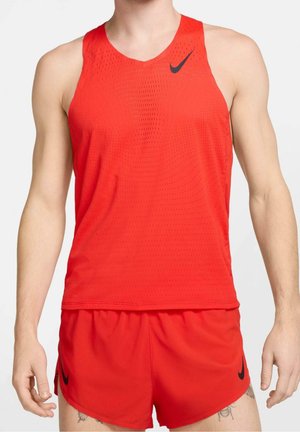 Atleta masculino con camiseta deportiva sin mangas roja y pantalones cortos con logotipos negros de Nike sobre un fondo liso.