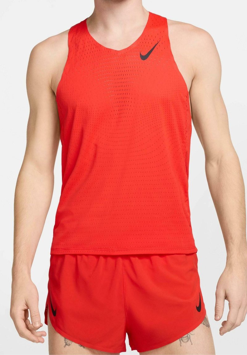 Athlète masculin portant un maillot de sport rouge sans manches et un short avec des logos Nike noirs sur un fond uni.