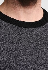 Grauer strukturierter Pullover mit einem schwarzen gerippten Kragen, der ein dezentes Muster im gesamten Stoff aufweist. Nahaufnahme des Kragenbereichs.