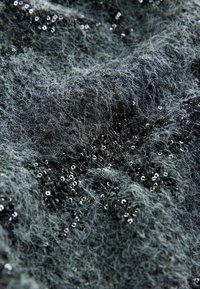 Gros plan d'un tissu gris texturé avec des fibres lâches et des paillettes noires dispersées de façon inégale sur la surface.