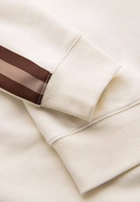 Sweatshirt crème avec des poignets côtelés ; présente un accent marron et rayé sur la manche. Texture douce avec des détails de couture soignée.