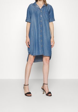 Mujer con un vestido camisero suelto de mezclilla azul, de manga corta y aberturas laterales, combinado con sandalias negras de tacón con tiras alrededor del tobillo.