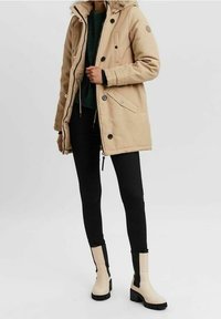 Parka beige con cappuccio peloso, con bottoni neri e tasche, indossata sopra un maglione verde scuro e leggings neri, abbinata a stivaletti beige alla caviglia.