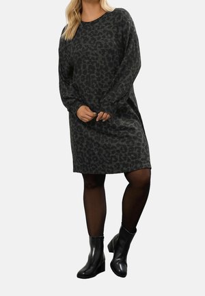 Femme portant une robe à manches longues, gris foncé à imprimé léopard, avec des collants noirs et des bottines noires, debout les mains jointes.