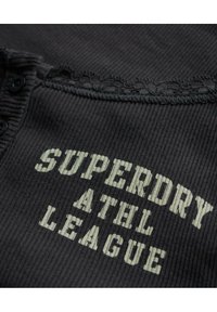 Zwarte ribbelstof met een gegolfde rand en knoopdetail. Bevat de tekst "SUPERDRY ATHL LEAGUE" in lichtgrijs.