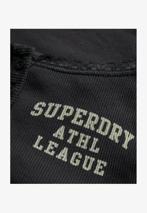 Zwarte ribbelstof met een gegolfde rand en knoopdetail. Bevat de tekst "SUPERDRY ATHL LEAGUE" in lichtgrijs.