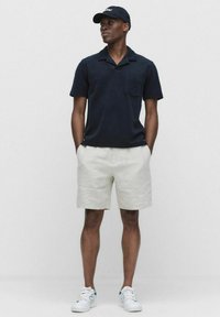 Marineblauwe polo met korte mouwen en zak, gecombineerd met lichtbeige shorts en witte sneakers met groene accenten.