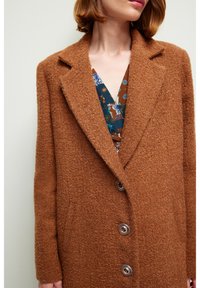 Femme portant un manteau en laine marron texturé par-dessus un chemisier à col en V bleu et marron à motif, cadré au-dessous des épaules.