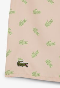 Beiger Stoff mit kleinen grünen Krokodilmotiven und einem größeren, gestickten grünen Krokodil-Logo nahe dem unteren Rand.