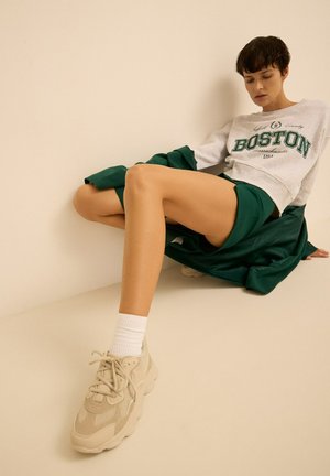 Felpa grigia chiara con la scritta "BOSTON", pantaloni corti verdi e sneakers chunky beige. Calzini bianchi alla caviglia completano l'outfit. Sfondo semplice.