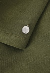 Material de culoare verde măsliniu, cu o țesătură texturată, având un eticheta metalică argintie cu un design floral atașată printr-un mic inel.