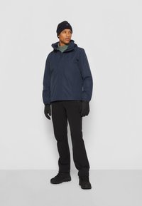 Veste étanche navy avec capuche, associée à un pantalon noir, des gants noirs et des bottes. Texture lisse ; design minimaliste ; détails pratiques.