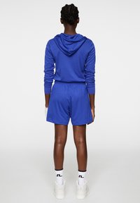 Sininen urheilupaita hupulla, yhdistettynä vastaaviin shortsit. Omistaa venyvän kankaan ja joustavan vyötärön. Valkoiset korkeakärkiset sneakerit täydentävät asua.