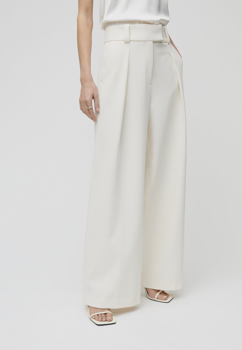 Weite Beinform-Hosen in Creme, mit einem strukturierten Bund, seitlichen Taschen und einer glatten Textur. Kombiniert mit minimalistischen weißen Sandalen.